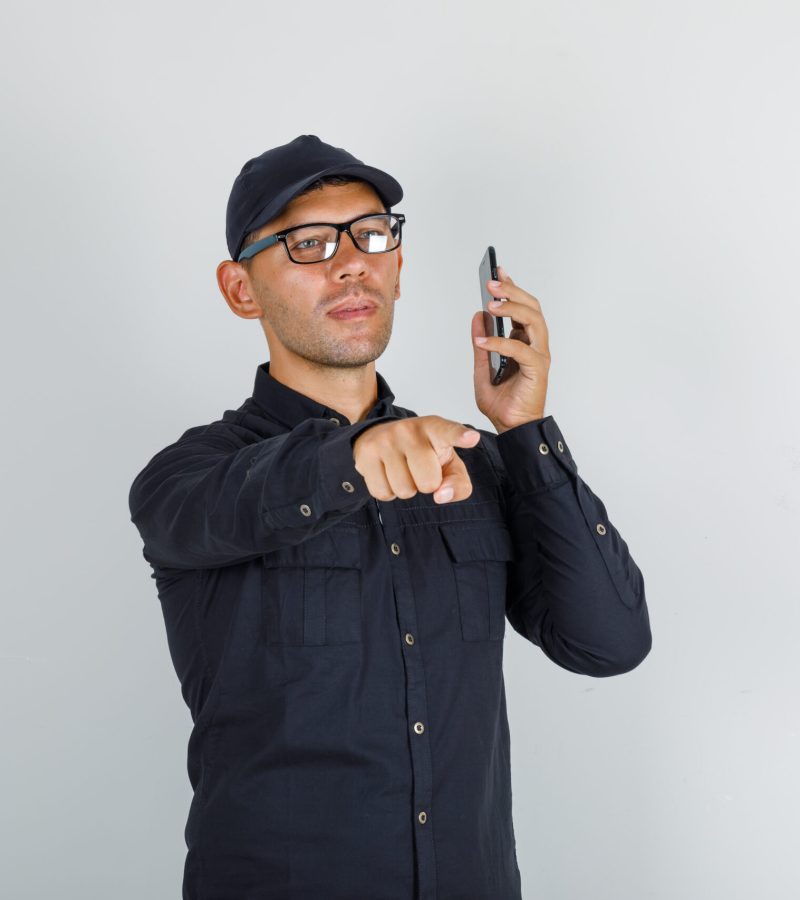 Young man pointing finger to camera with smartphone in black shirt, cap, glasses , front view.